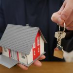 Mão segurando casa em miniatura e chaves, representando a compra de imóvel por consórcio